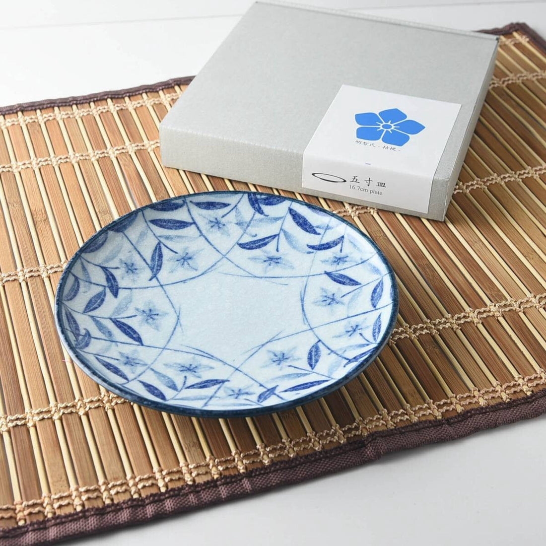 A small blue-and-white plate painted with balloon flowers, beside its gift box on a bamboo mat