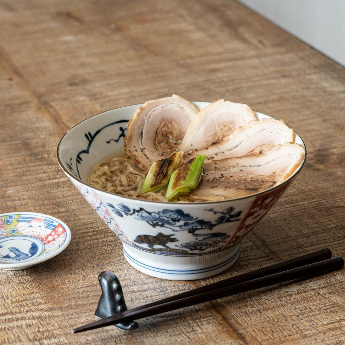 A bowl of ramen in an indigo-and-red pine and plum donburi bowl, with chopsticks, on a wooden table