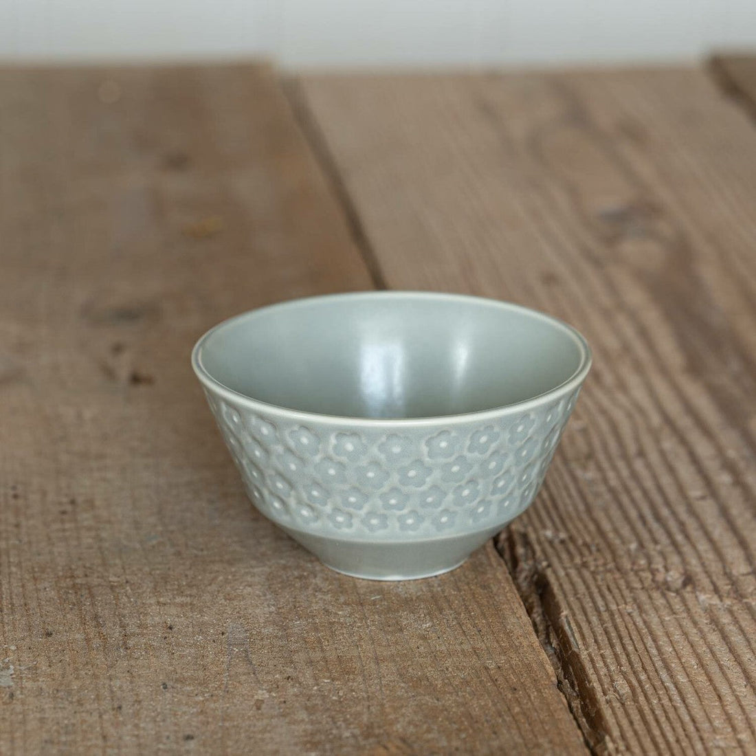 A matte gray porcelain rice bowl with pressed flowers around the outside, on a worn wooden table